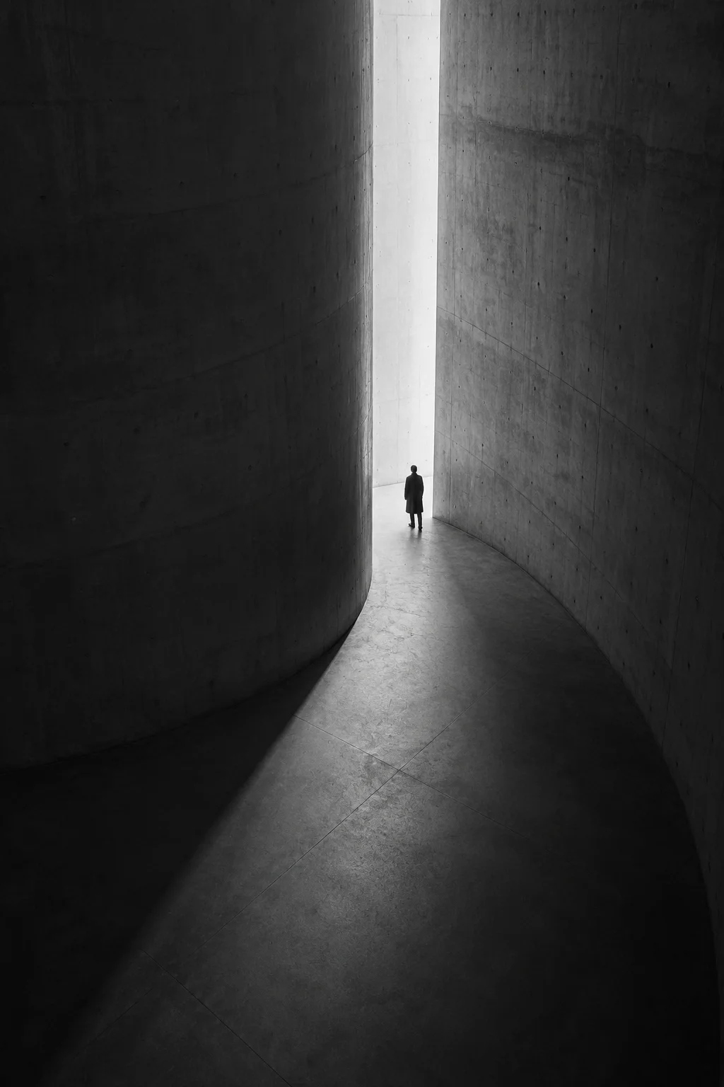 A solitary figure framed by curved concrete walls and deep shadows in black and white